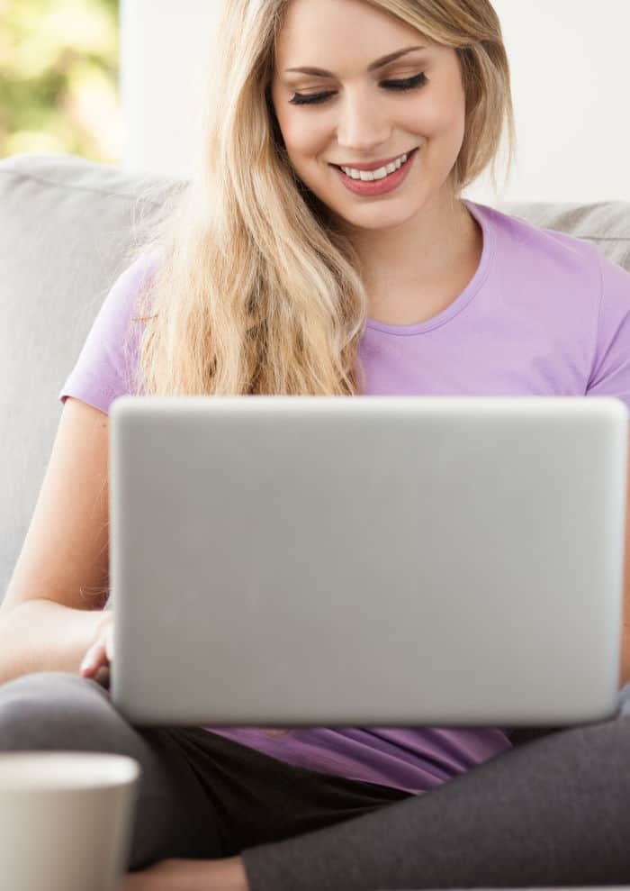 A woman sits facing forward smiling and working on a laptop from her couch.