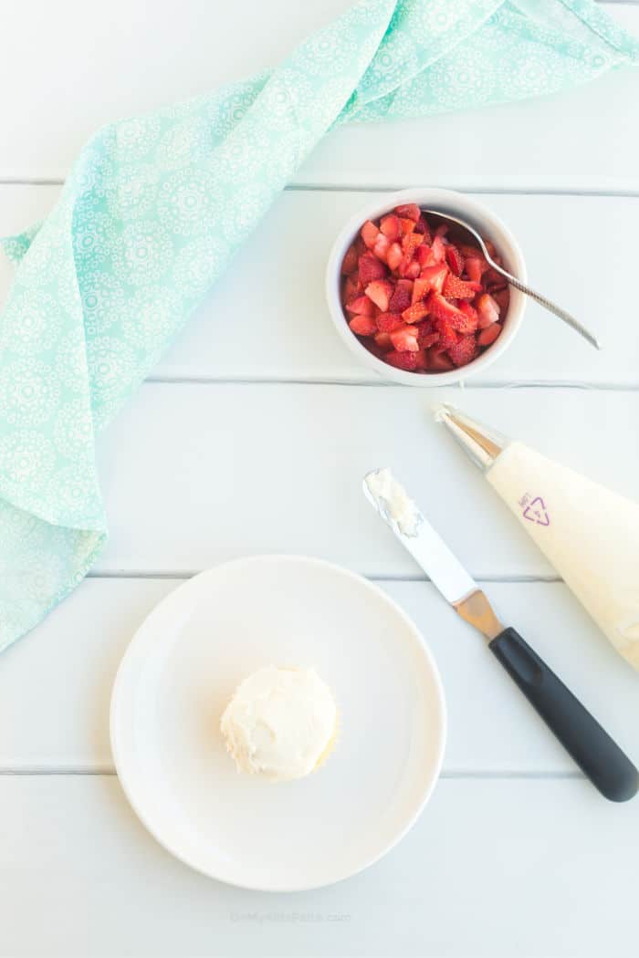 A cupcake bing frosting with a piping bag and spatula from overhead, and a bowl of diced strawberries.