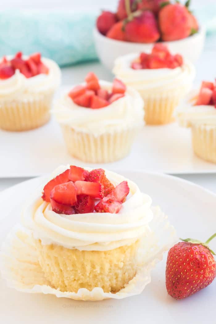 A close-up of a cupcake with frosting and fresh diced strawberries being unwrapped, with more cupcakes on a plate behind.