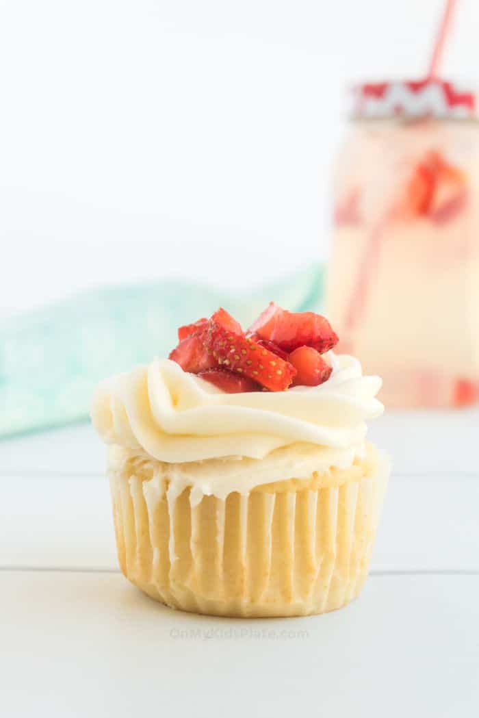 A close up of a cupcake with frosting and fresh strawberry topping, with strawberry lemonade in the background.
