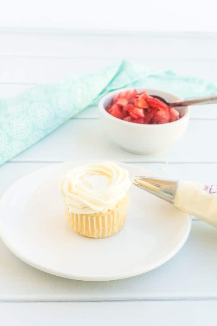 A cupcake being frosted with a piping bag in a swirl with a bowl of fresh strawberries in the background.