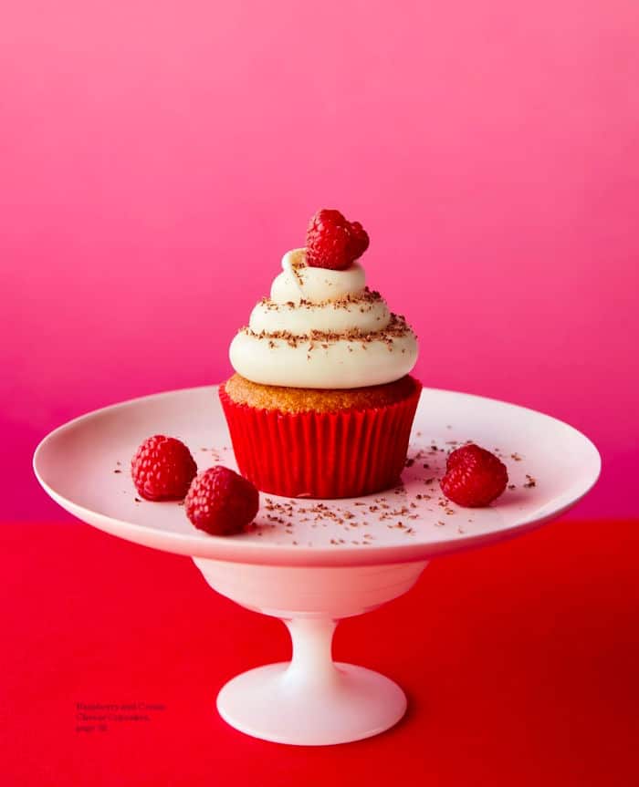 A raspberry cupcake sits on a cakestand with a raspberry on top and raspberries around the plate