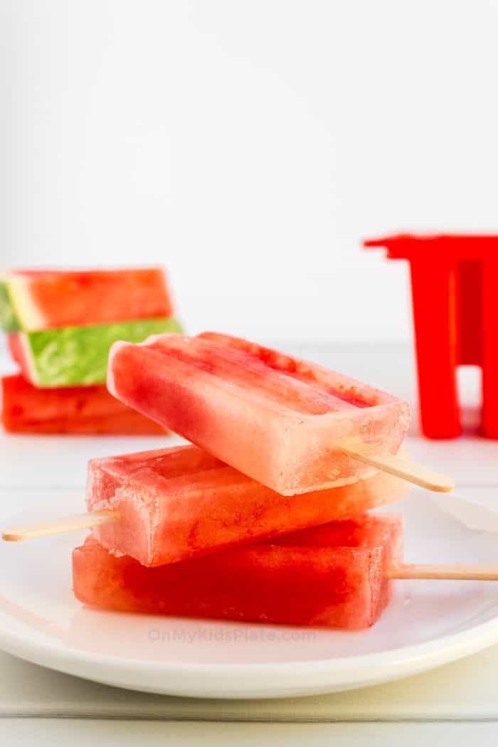 Watermelon popsicles are stacked three high in the front. In the background is slices of fresh watermelon and a popsicle mold.