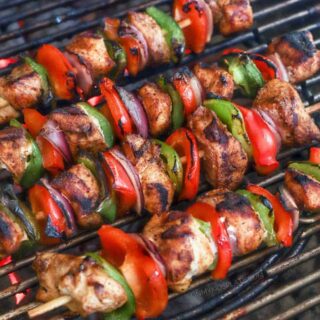 A close up view of chicken, peppers and onions threaded on skewers cooking on a grill.