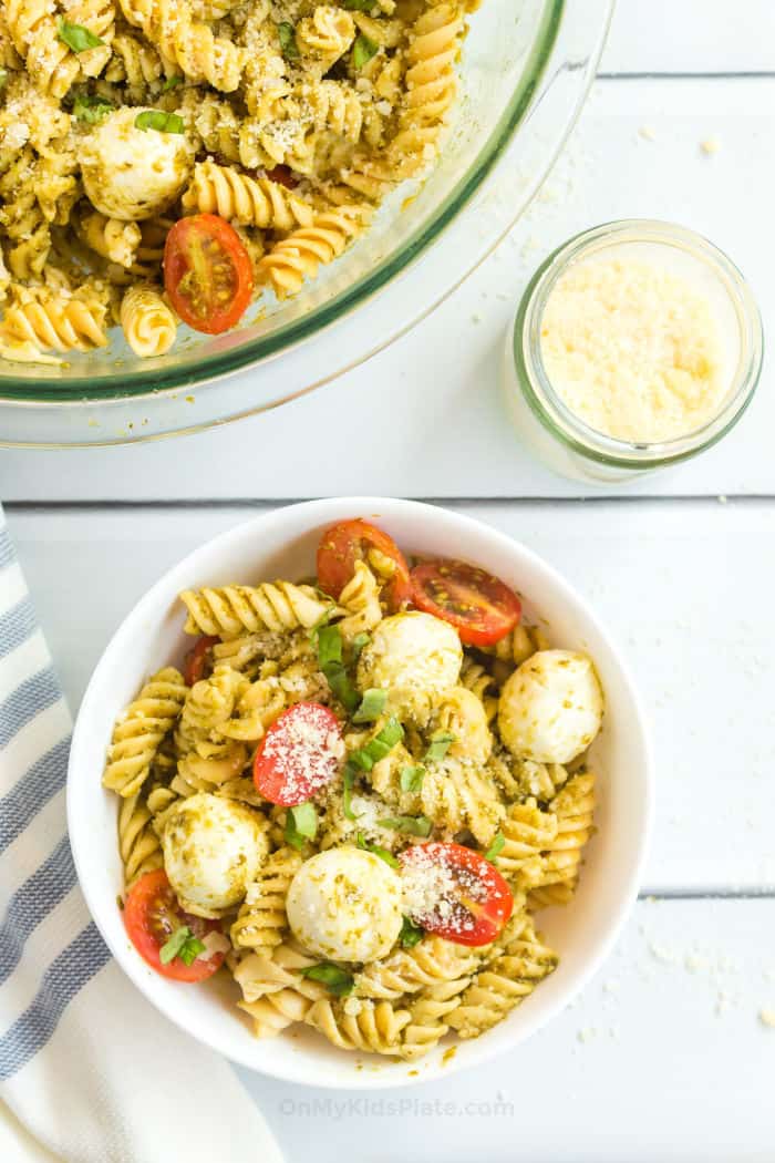 Pesto Pasta salad sits mixed in a small white bowl next to a small container of parmesan cheese and a larger serving bowl of pasta.