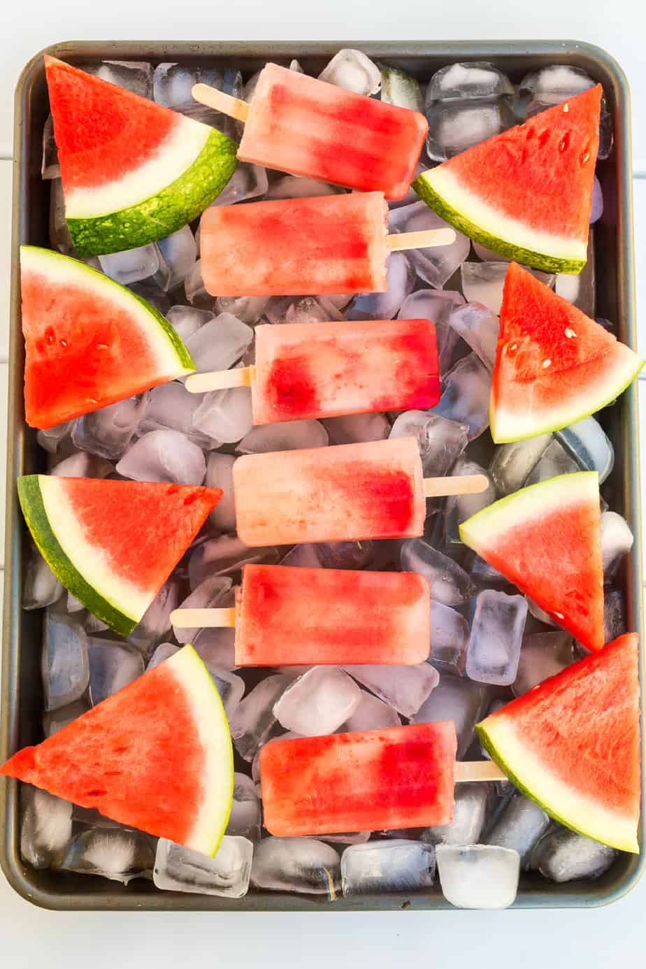 Watermelon popsicles and slices of fresh watermelon on a tray covered in ice from overhead.