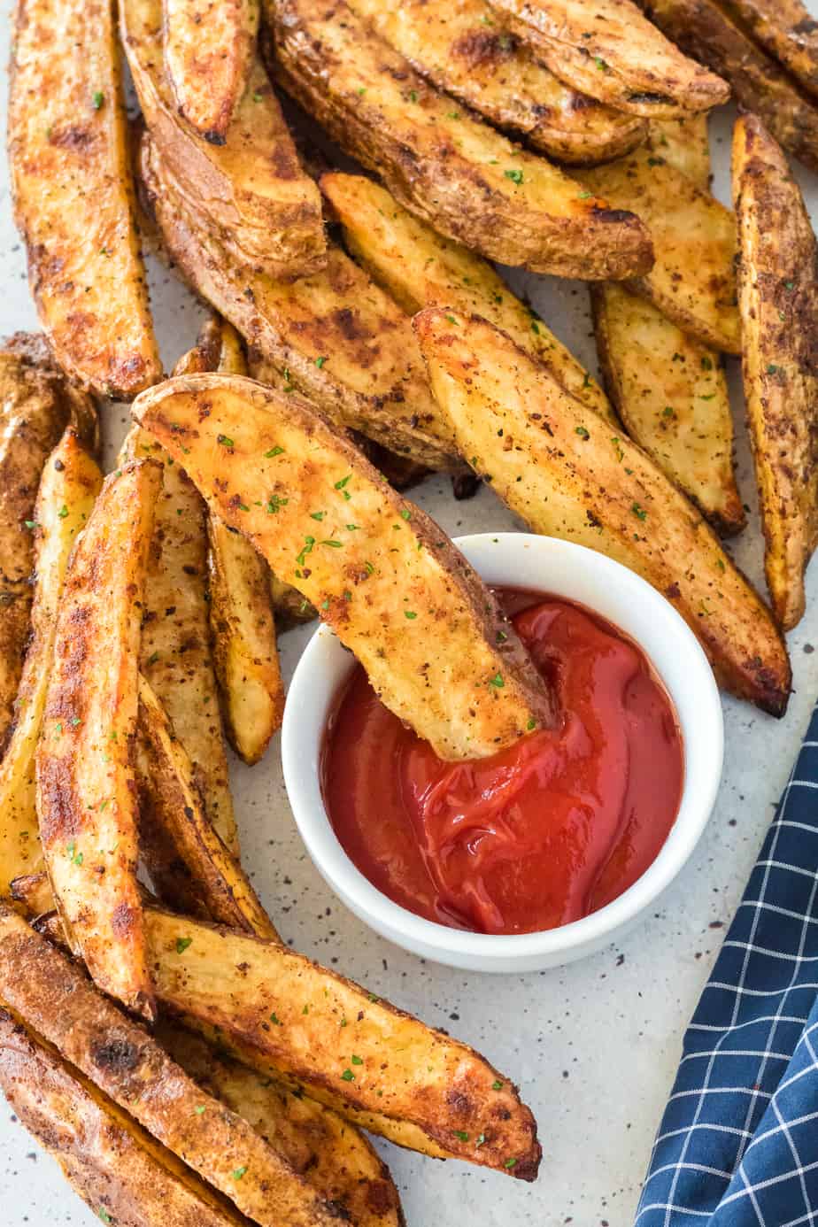 Crispy brown potato wedges piled on a serving platter close up with one potato slice being dipped in a small bowl of ketchup