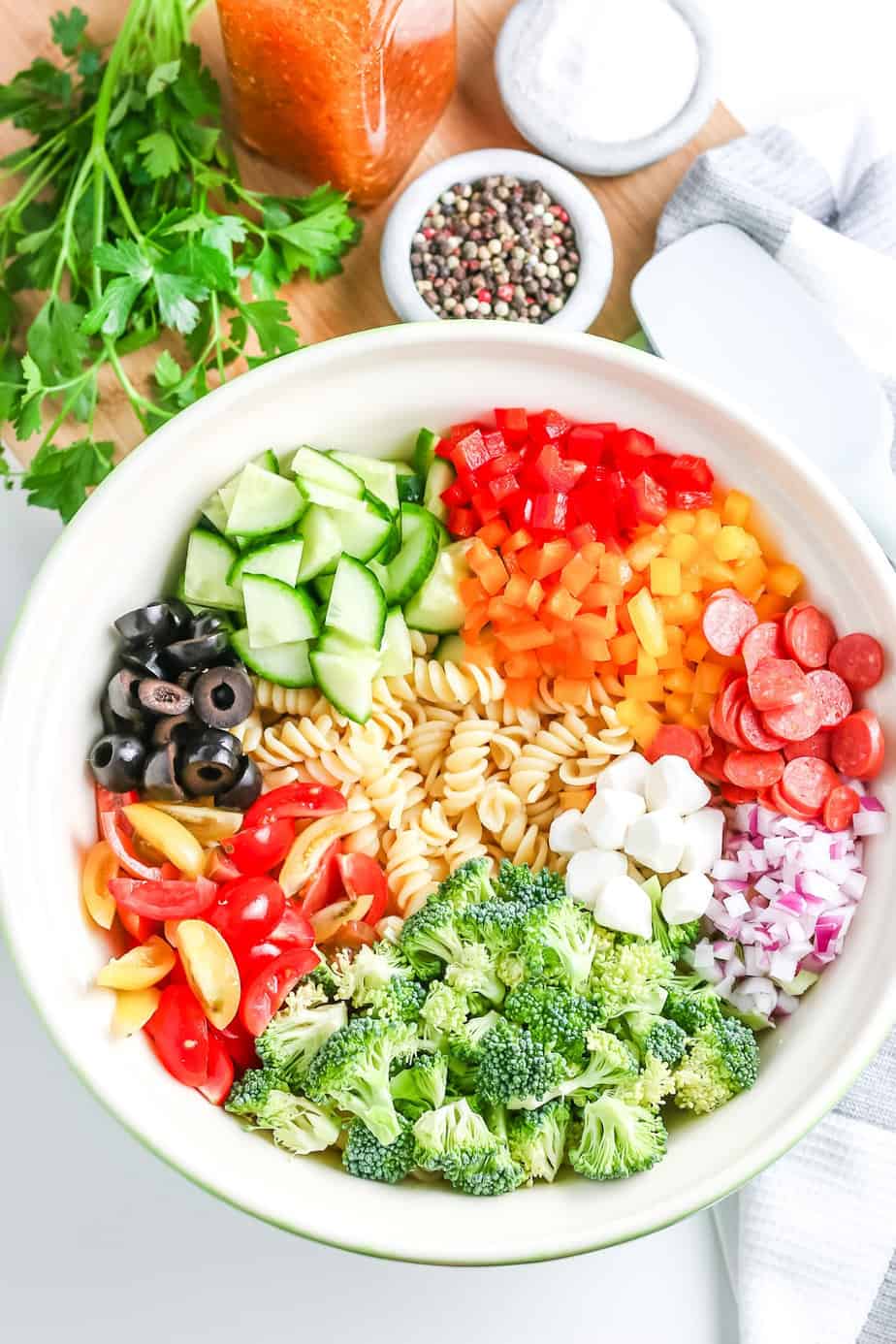 A bowl of pasta salad in a bowl from overhead with the vegetables, cheese and pepperoni chopped before mixing with more fresh ingredients on a cutting board nearby.