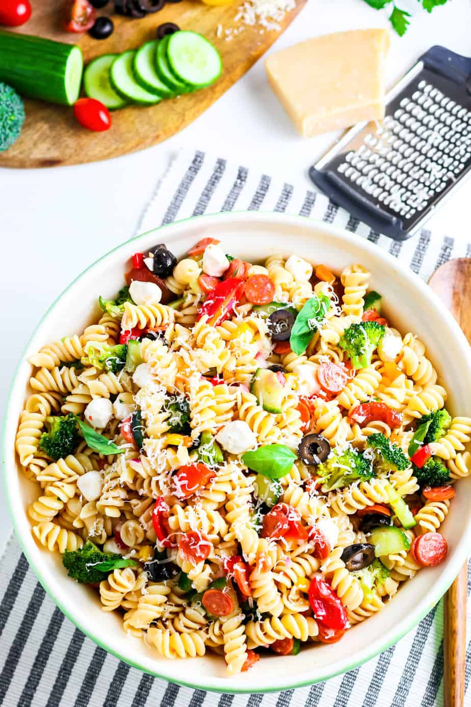Italian pasta salad in a large serving bowl with a cutting board with vegetables and parmesan cheese and a grater near the bowl.