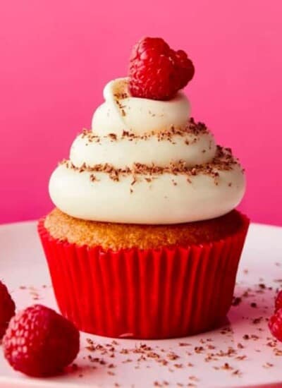 A raspberry cream cheese cupcake on a plate surrounded by scattered fresh raspberries
