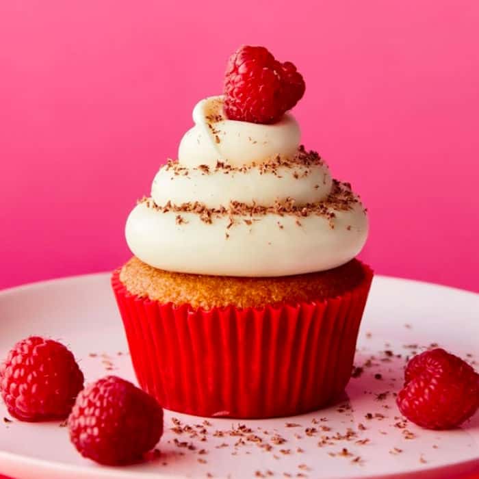 A raspberry cream cheese cupcake on a plate surrounded by scattered fresh raspberries