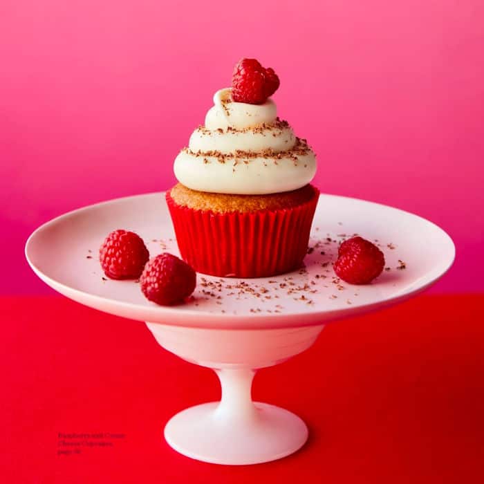A raspberry cream cheese cupcakes sits on a cake plate surrounded by fresh raspberries and chocolate shavings.