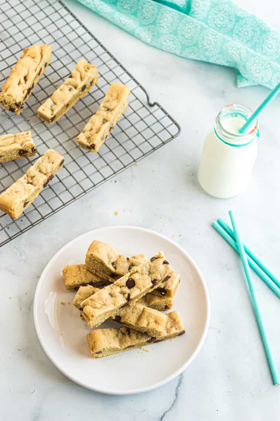 Chocolate chip cookie sticks on a serving plate next to a glass of milk, with a few cookies in the background on a cooling rack.