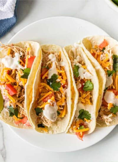 four tacos on a plate loaded with shredded chicken, cheese, sour cream and cilantro