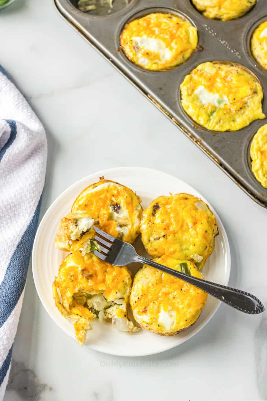 Egg bites on a plate with a fork, a muffin pan with baked omelets insider to the side.