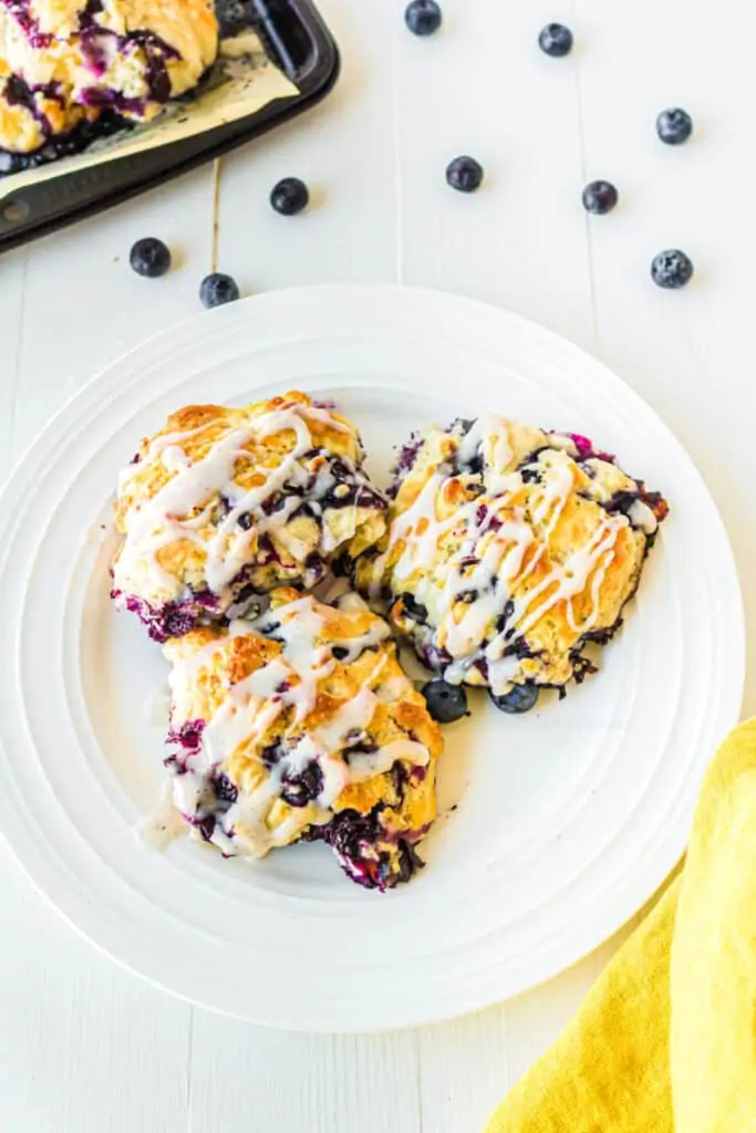 Three blueberry biscuits with glaze on a plate from overhead next to a pan of biscuits