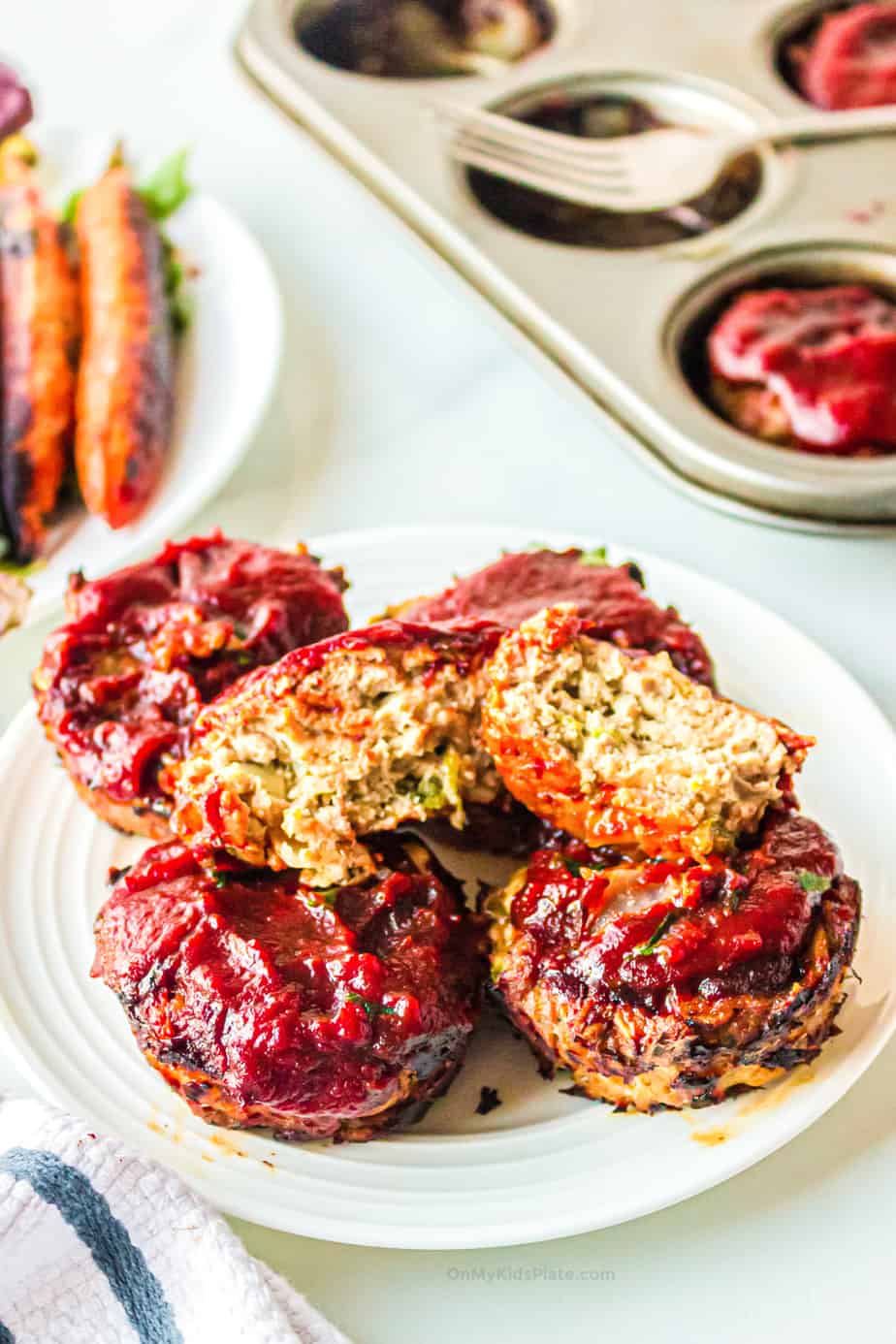 Stack of mini turkey meatloaf muffins on a plate with the muffin on top broken open. A muffin pan and roasted carrots are in the background.