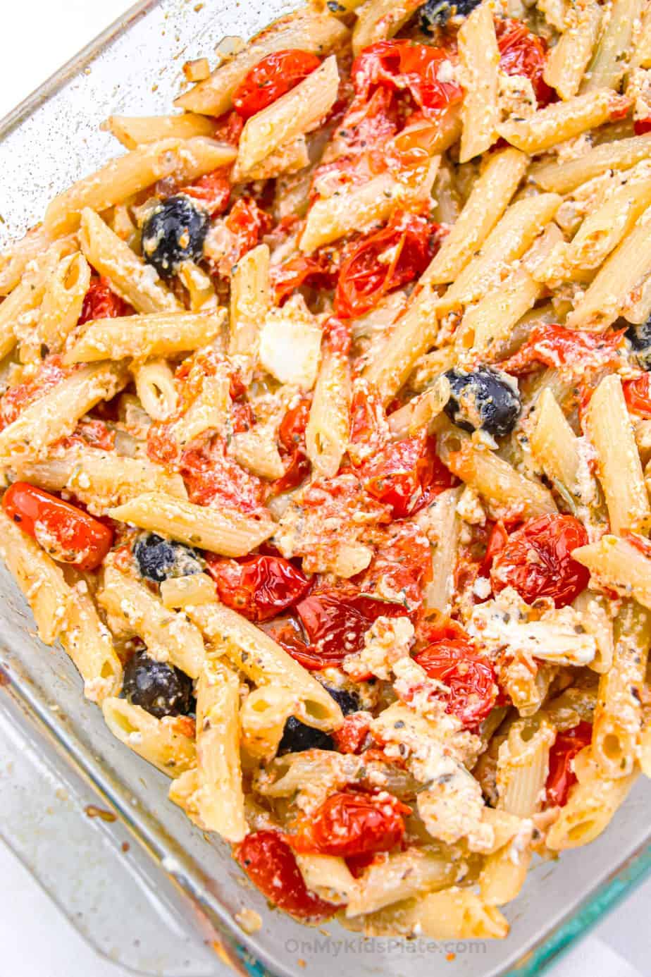 Penne pasta with roasted tomatoes, olives and feta cheese mixed in a casserole dish.