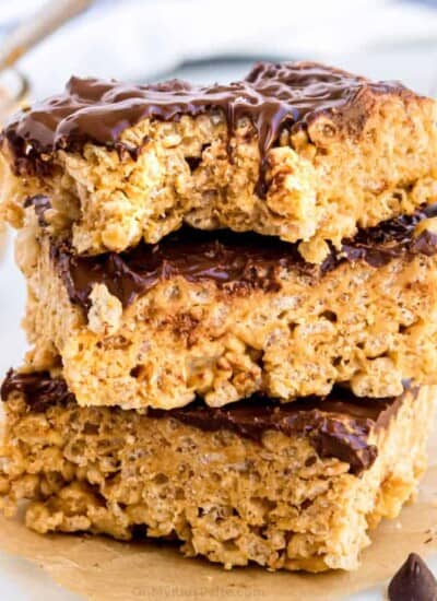 Stacked Rice Krispie treats covered in chocolate with peanut butter and chocolate chips in the background.