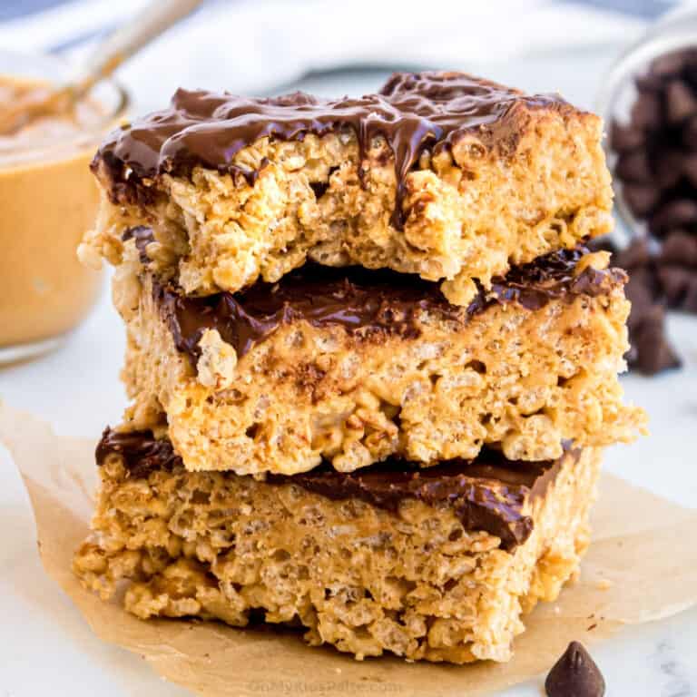 Stacked Rice Krispie treats covered in chocolate with peanut butter and chocolate chips in the background.