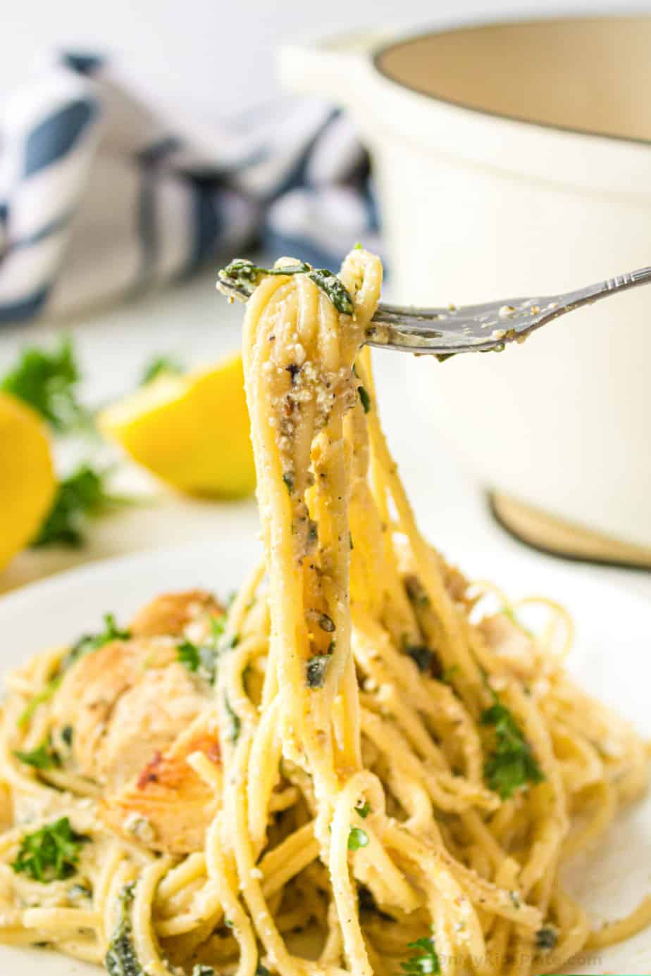 Pasta with chicken, cheese and herbs being pulled by a fork from a plate with lemon in the background
