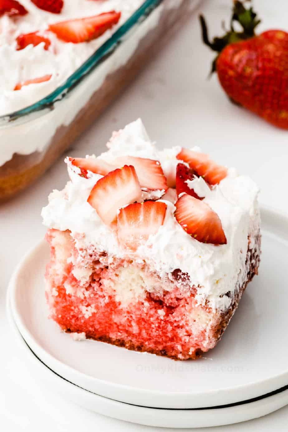 A slice of red and white cake topped with strawberries on a plate next to a pan full of cake.