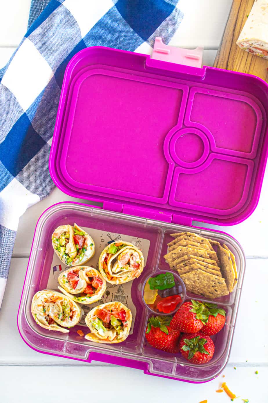 A child's lunchbox full of vegetable cheese pinwheels, strawberries, crackers and gummy bears.