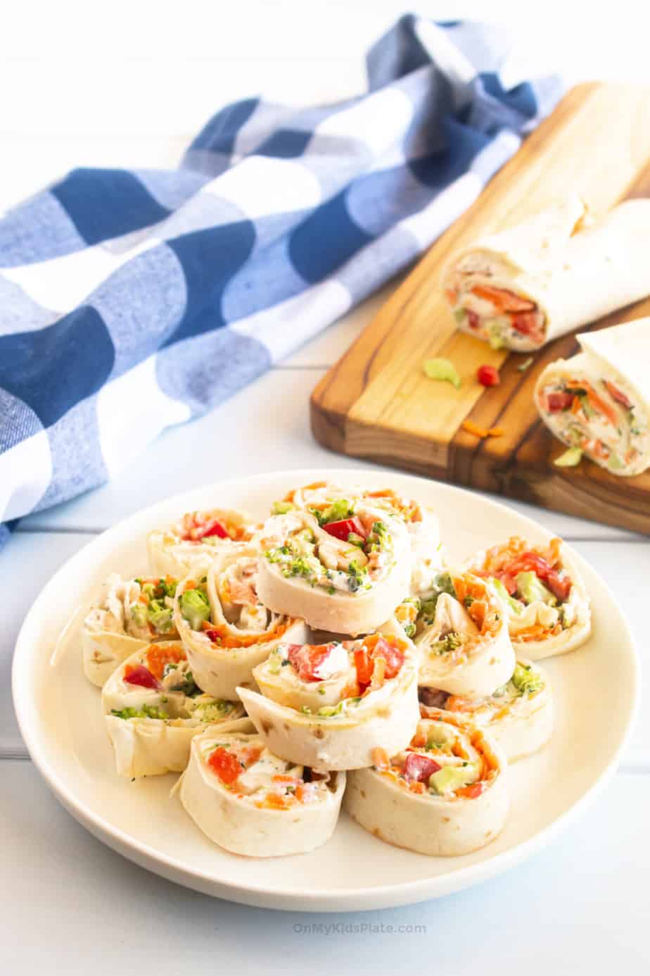 Vegetable cream cheese tortilla pinwheels sliced and stacked on a plate with a cutting board in the background