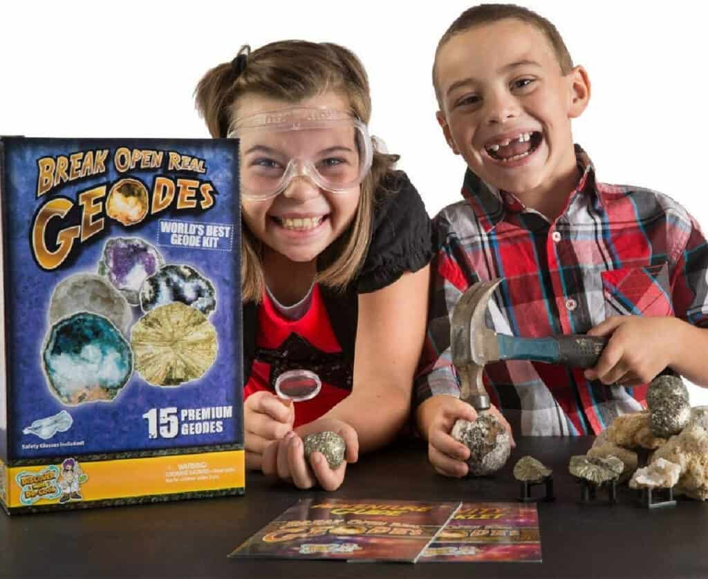 Kids wearing goggles holding geodes and a hammer next to kit box.