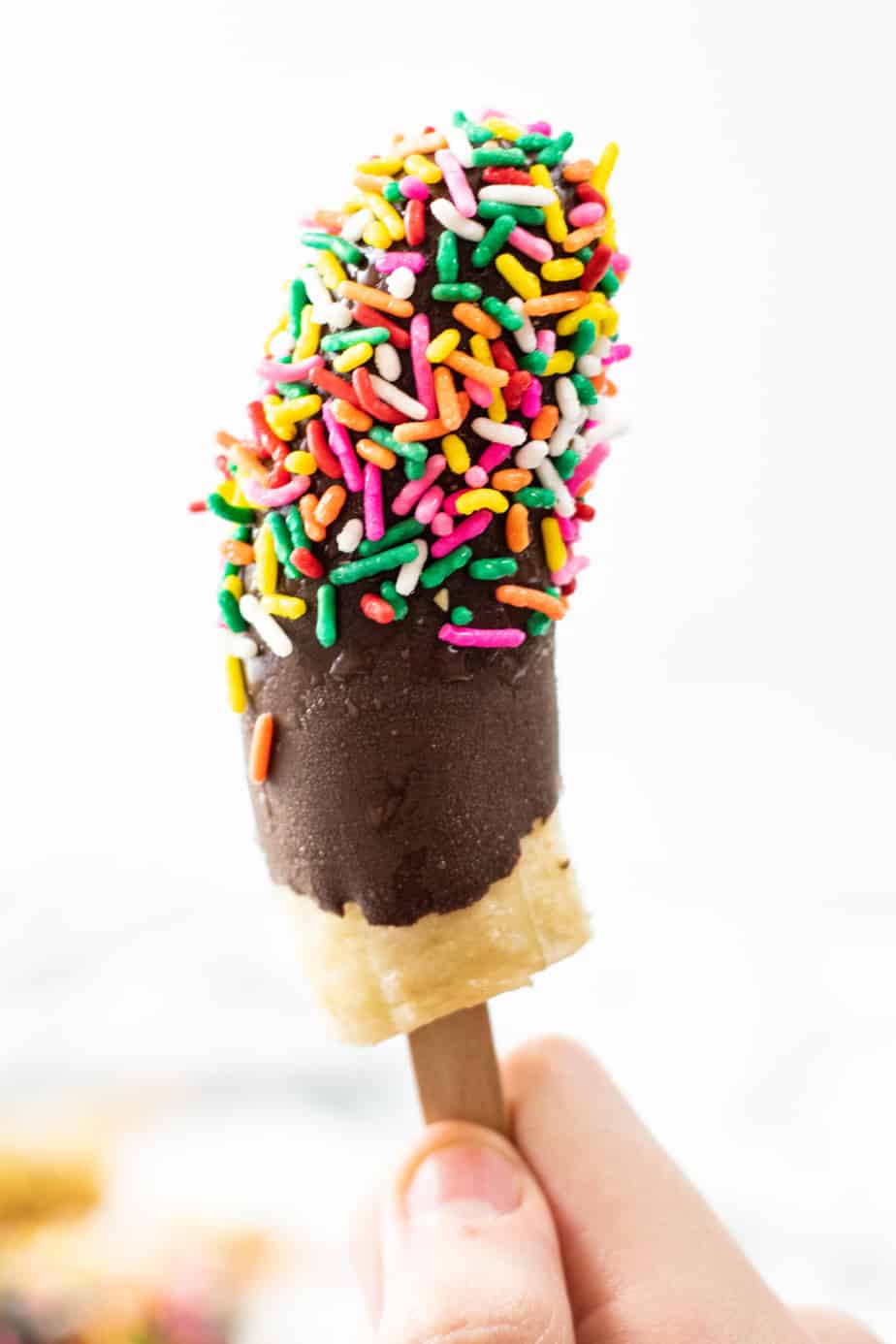 Frozen banana pop covered in chocolate and sprinkles being held in the air close up.