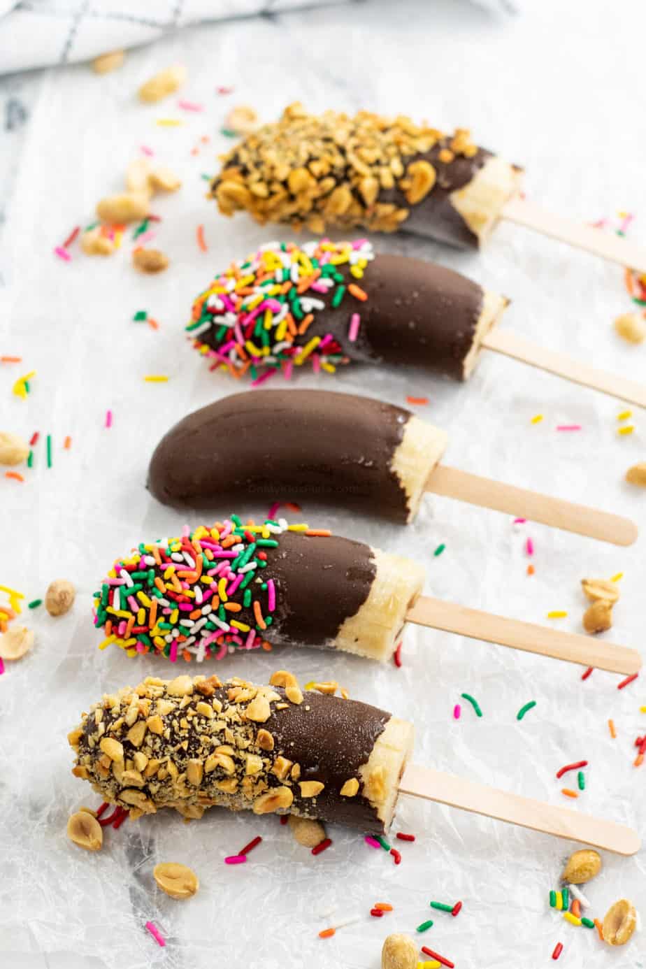 Frozen banana popsicles lined up on wax paper topped with nuts and sprinkles