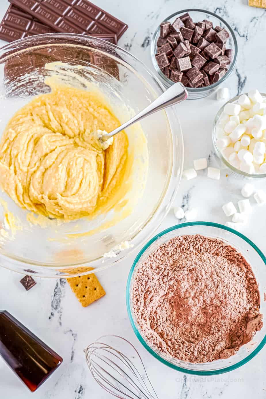 Two bowls with wet and dry cookie dough mixtures ready to be mixed and other cookie ingredients such as chocolate and marshamallows in bowls nearby.