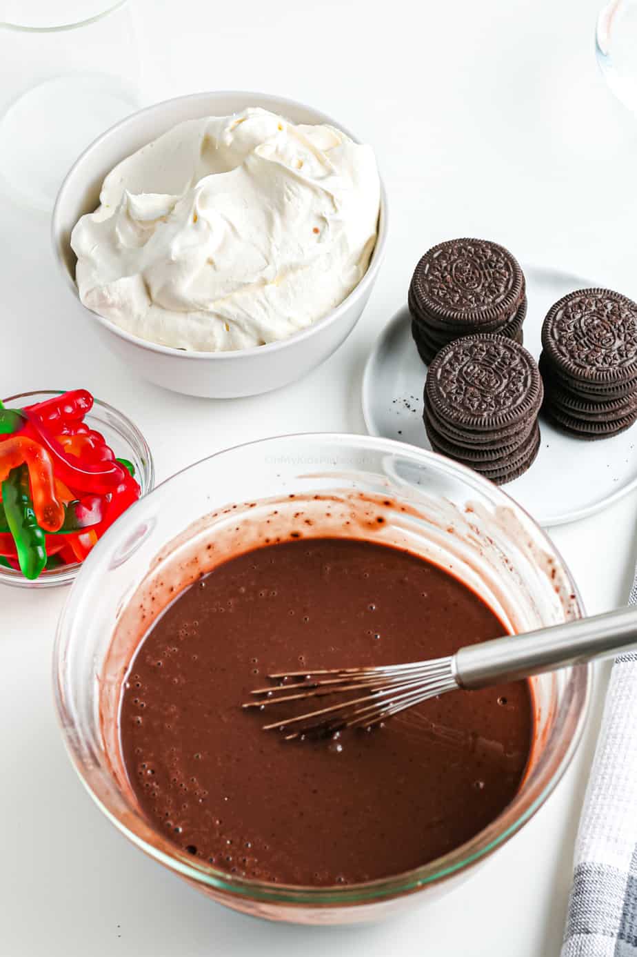 Chocolate pudding being mixed in a bowl with a whisk with whipped topping, chocolate Oreos and gummy worms in bowls nearby.