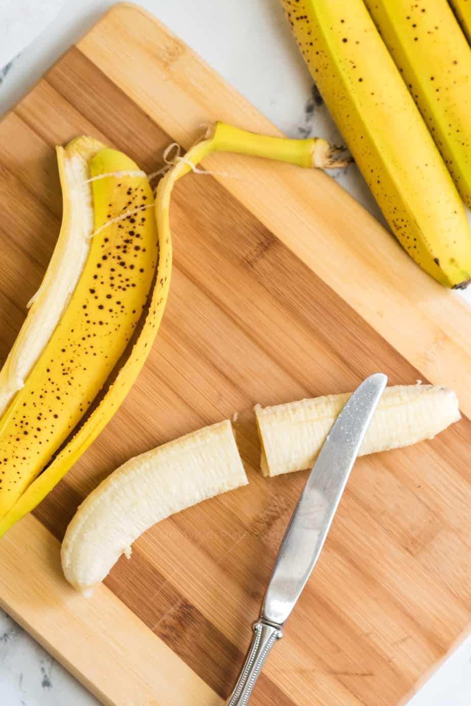 Banana being sliced in half with a knife on a cutting board next to the peel and more bananas