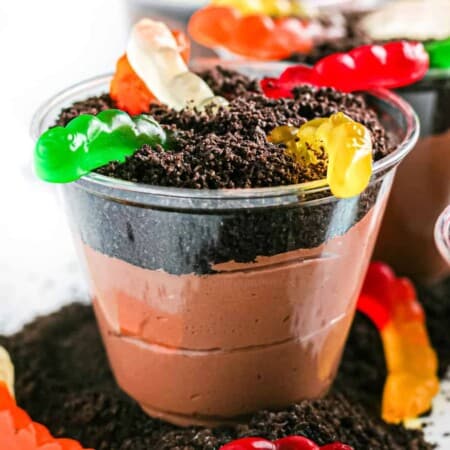 Up close of chocolate pudding in a clear cup layered with chocolate cookie crumbs and gummy worms to look like dirt.