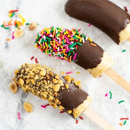 Three frozen bananas on popsicle sticks with nuts, sprinkles and chocolate