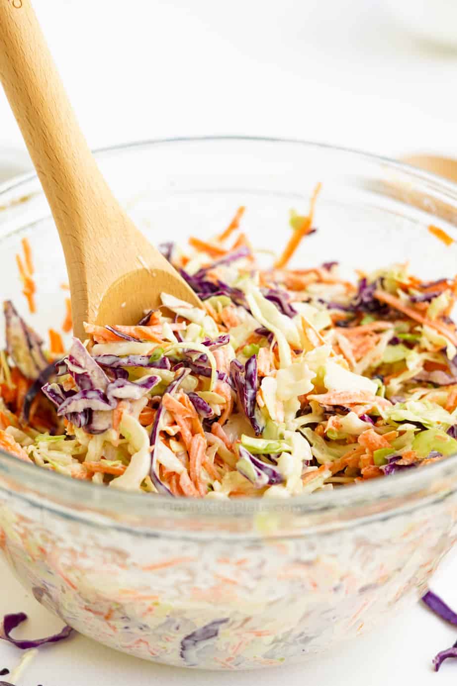 Mixing cabbage with dressing to make coleslaw in a large clear bowl from the side with a wooden spoon.