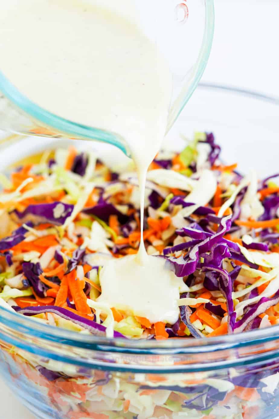 Pouring dressing over cabbage and carrot mixture in a clear bowl from the side.