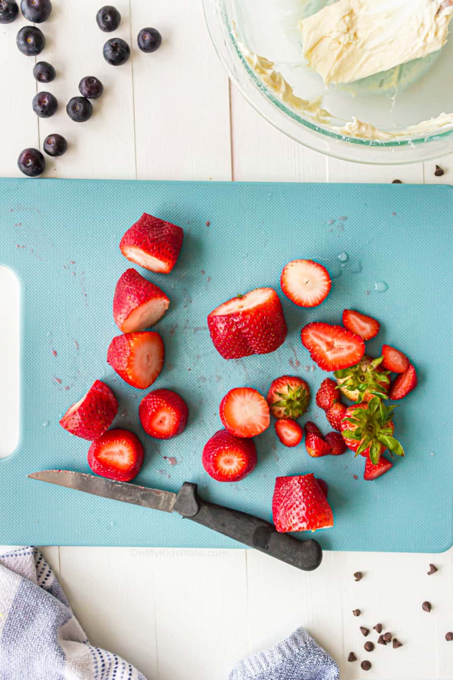 Strawberries with the tops and tips sliced off strewn across a cutting board with a small knife.
