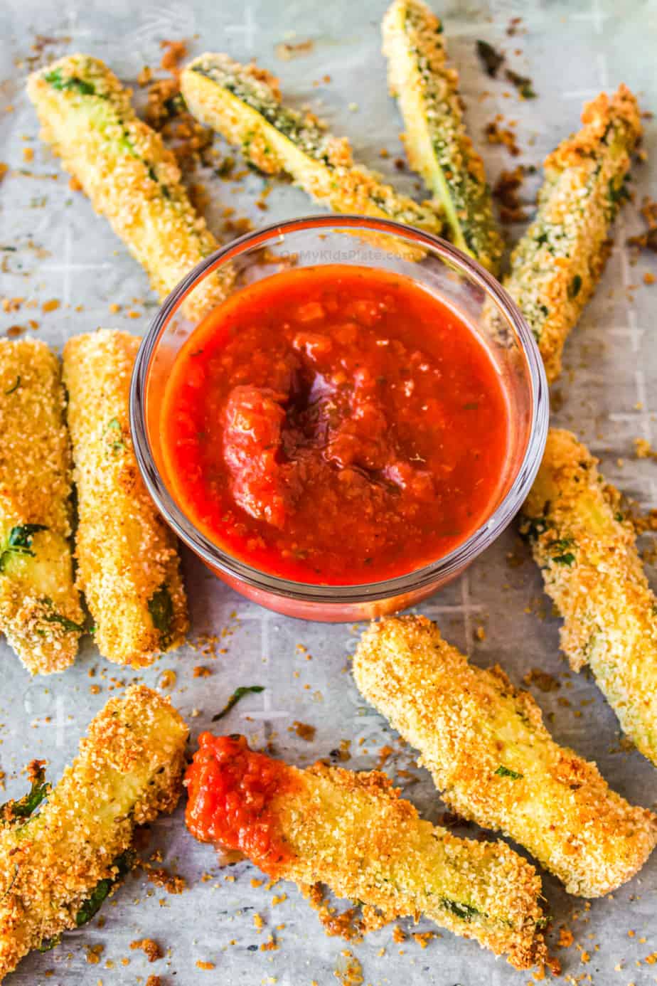 Golden brown zucchini fries next to marinara sauce on parchment paper. One fry is dipped in red marinara sauce.