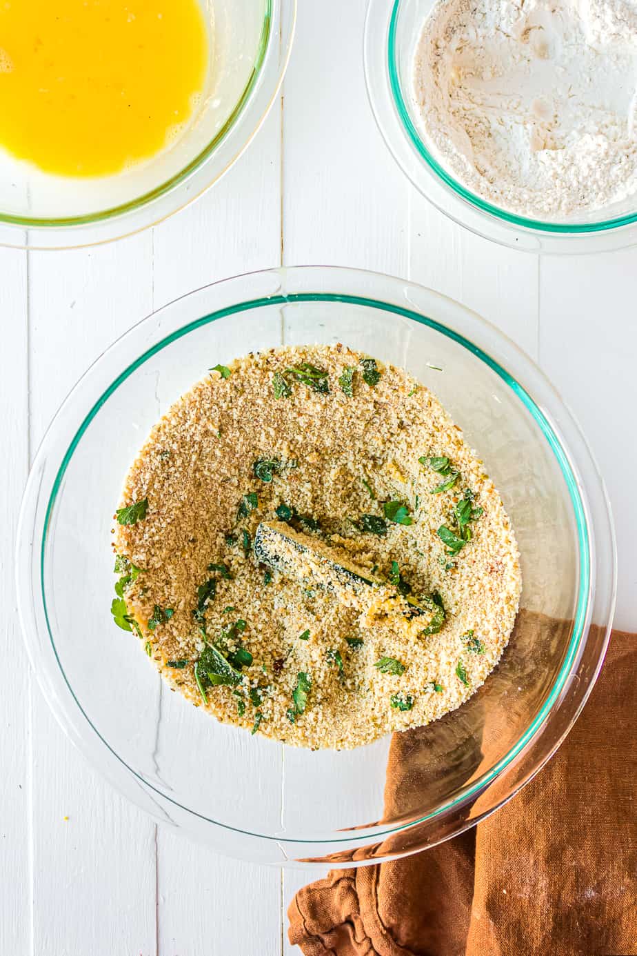 A slice of zucchini being coated in panko bread crumbs with herbs and parmesan cheese with a bowl of egg and a bowl of flour nearby from overhead.