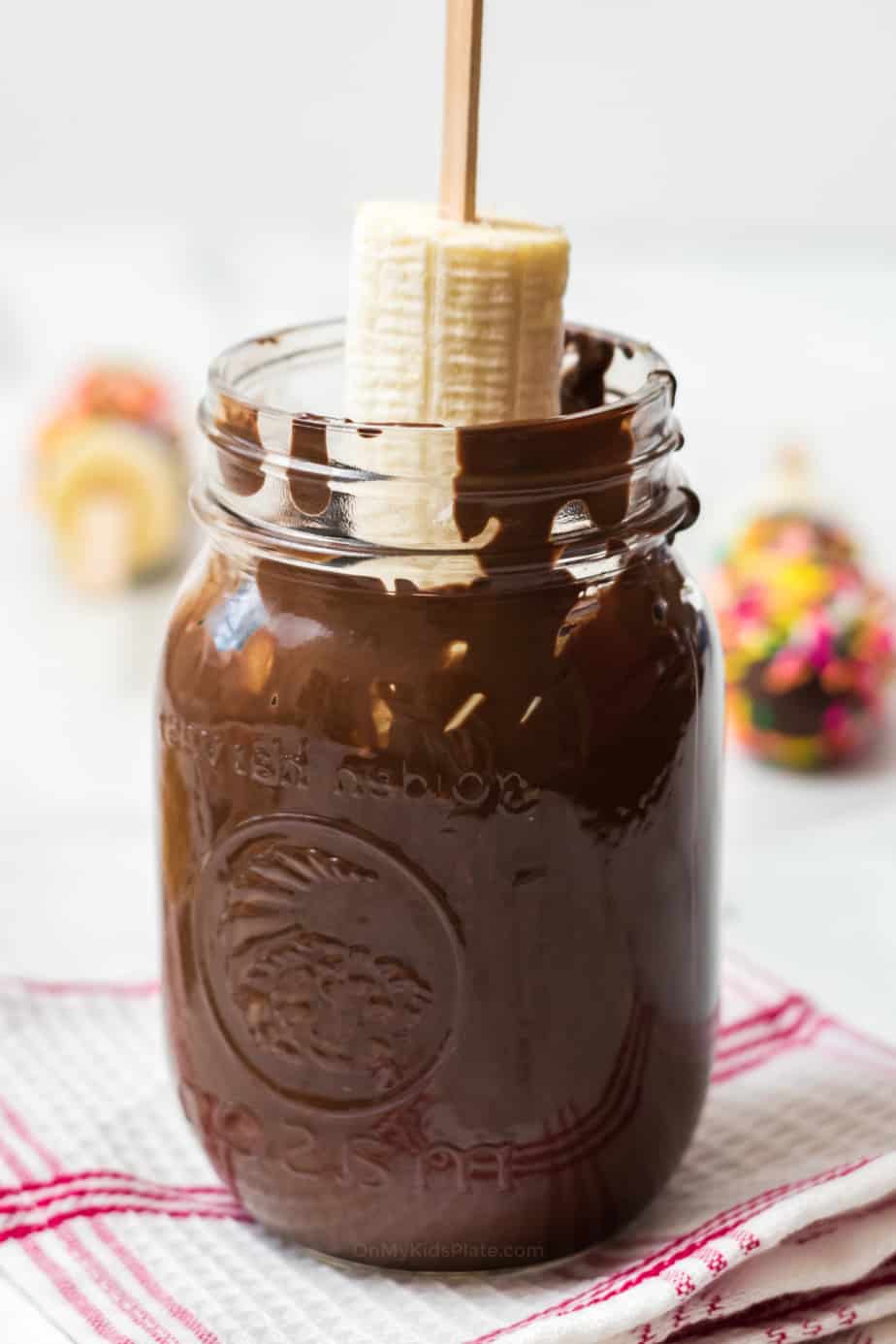 Banana on a stick being dipped in large jar of chocolate.