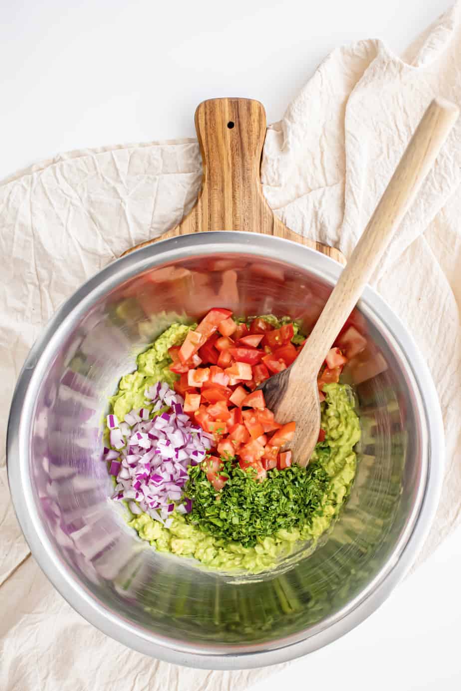 Mixing diced tomatoes, cilantro and purple onion into a large bowl of mashed avocado to make guacamole.