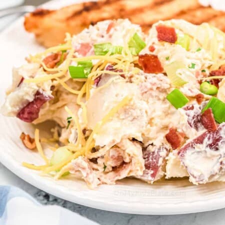 potato salad topped with cheese, green onions and bacon on a dinner plate with chicken behind