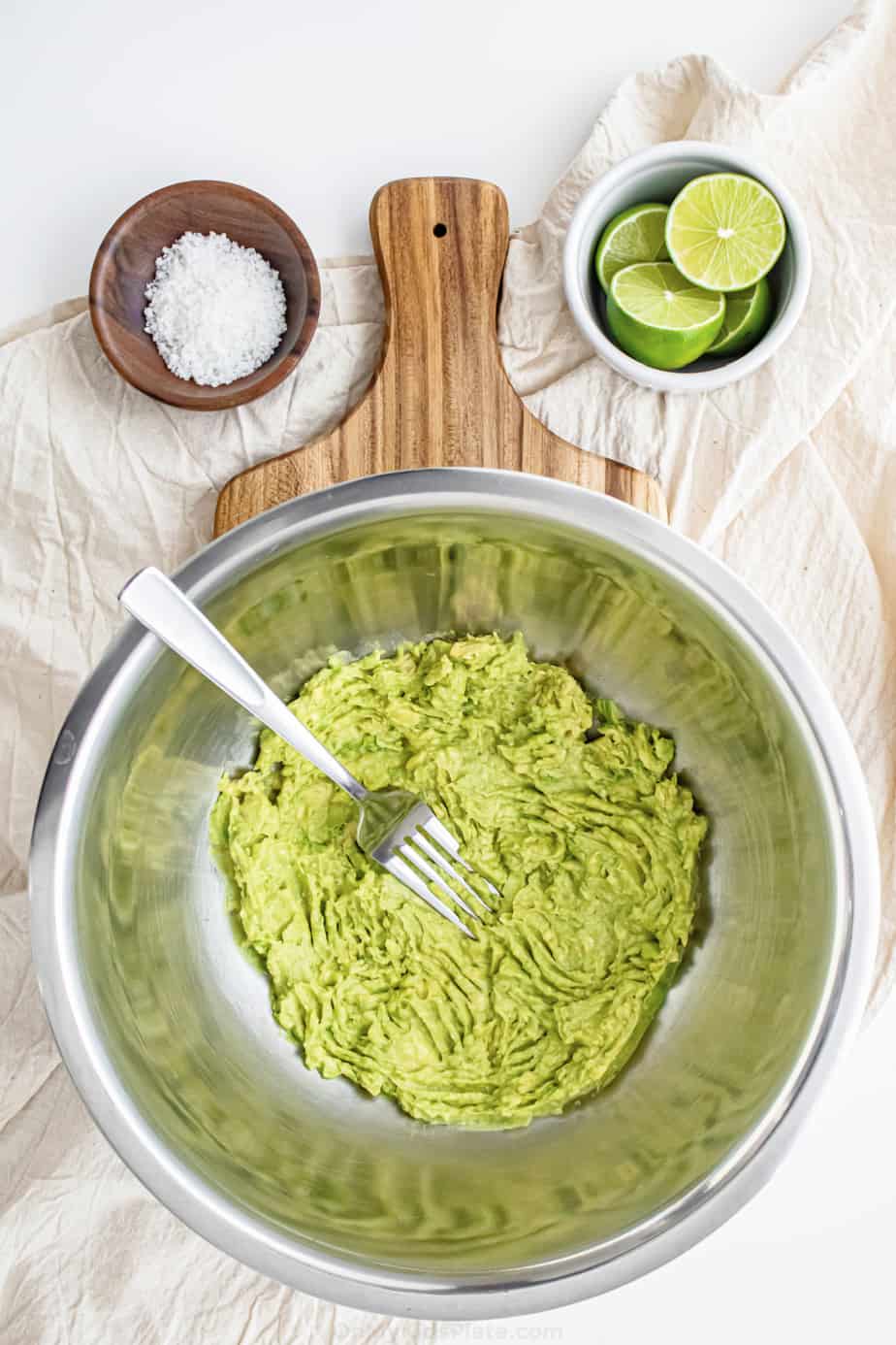 Mashing avocado in a large bowl with a fork with lime and salt on the side in bowls.