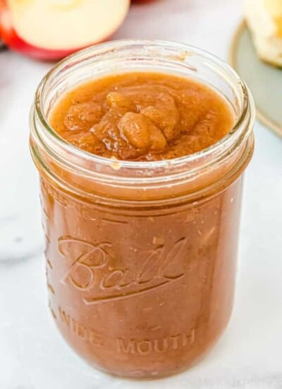 A mason jar of apple butter with apples and biscuits in the background