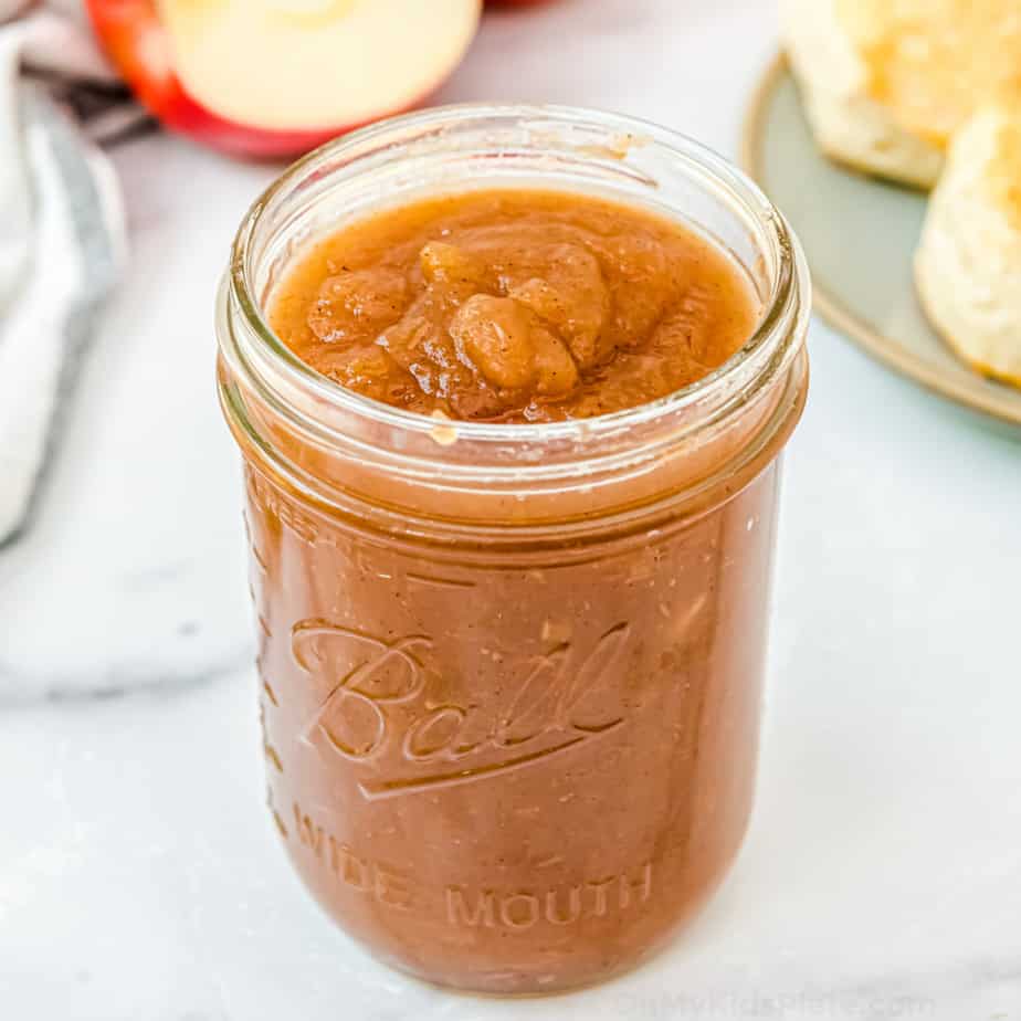 A mason jar of apple butter with apples and biscuits in the background