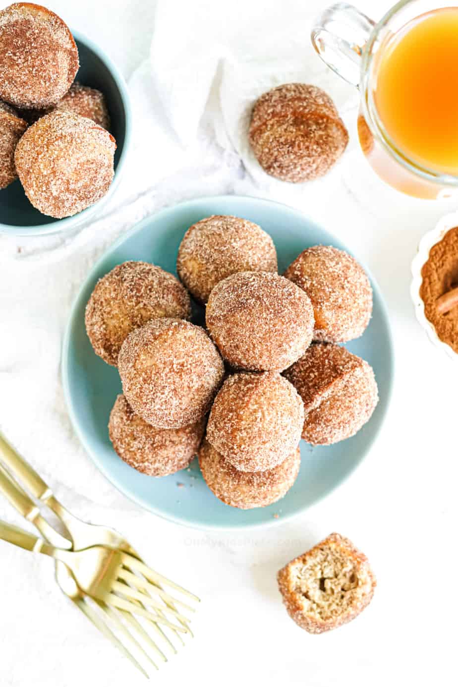 Plate of doughnut holes covered in cinnamon sugar from overhead with a glass of apple cider, forks and a second bowl of doughnut holes nearby on the table