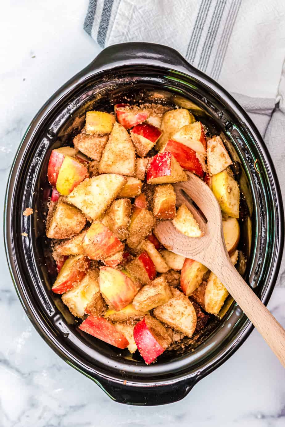 Sliced apples mixed with brown sugar and spices in a slow cooker with a spoon