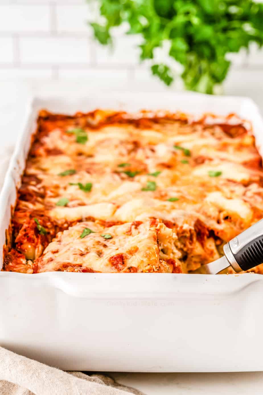 Baked ziti covered in a melted cheese in a pan with a spoon beginning to lift out a serving.